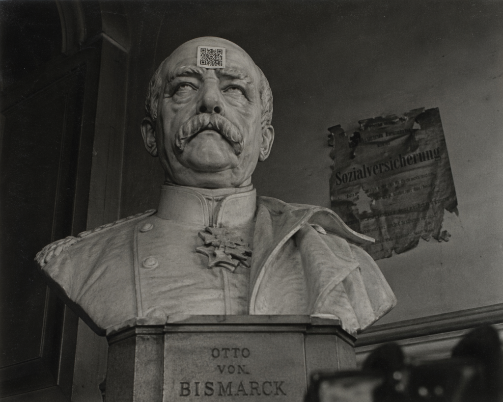 Büste von Otto von Bismarck mit einem QR-Code, im Hintergrund ein zerfetztes Zitat mit der Aufschrift "Sozialversicherung", historische Aufnahme